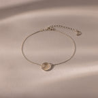 The Dainty Organic Fingerprint Bracelet in Sterling Silver features a round engraved charm, displayed on a beige textured background with soft shadows—an elegant, personalized jewelry piece.