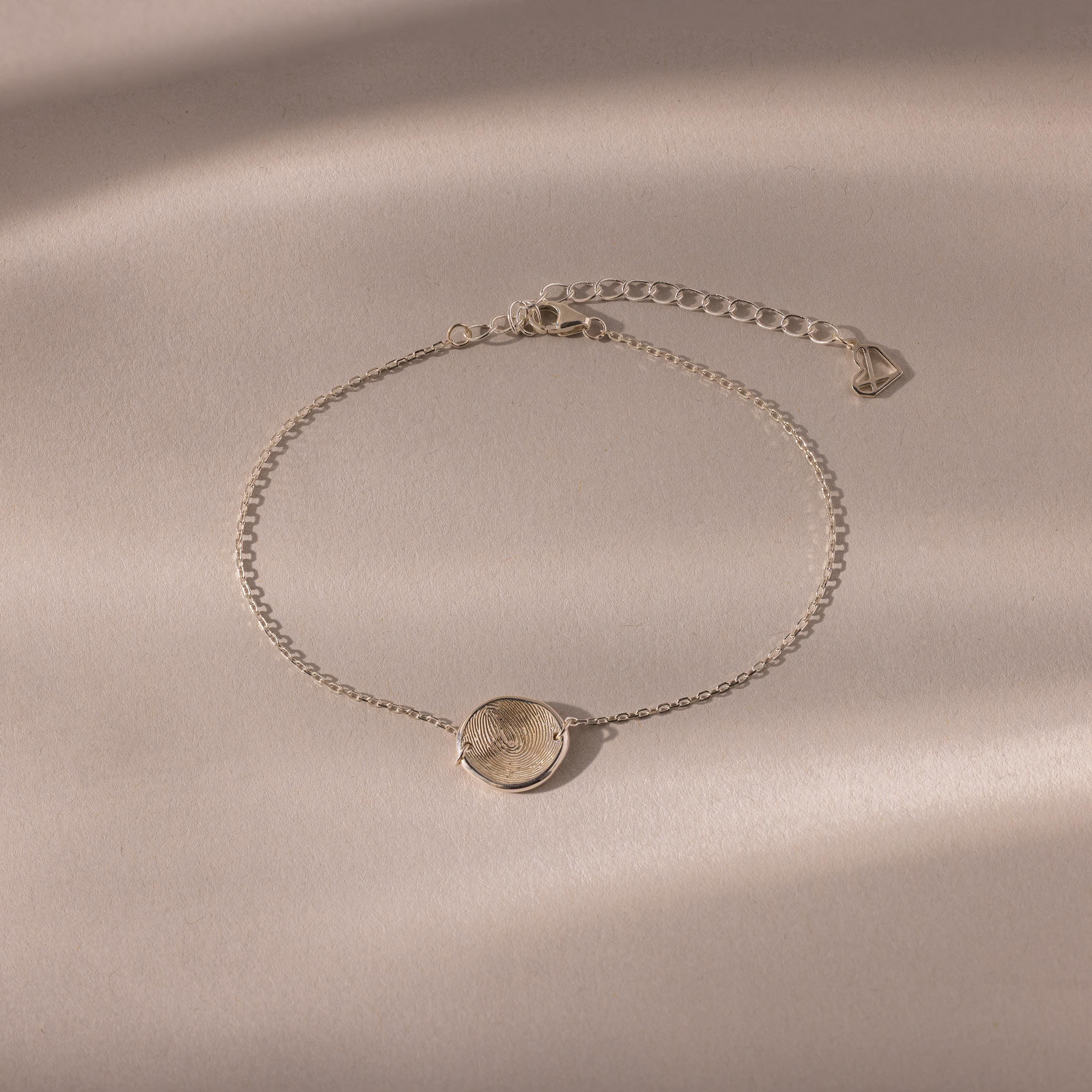 The Dainty Organic Fingerprint Bracelet in Sterling Silver features a round engraved charm, displayed on a beige textured background with soft shadows—an elegant, personalized jewelry piece.