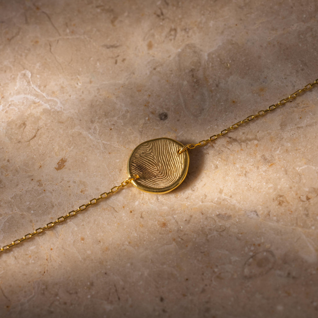 Dainty Organic Fingerprint Bracelet in 18K Gold with a round pendant featuring your unique design, displayed on a stone surface—a beautiful personalized memorial gift.