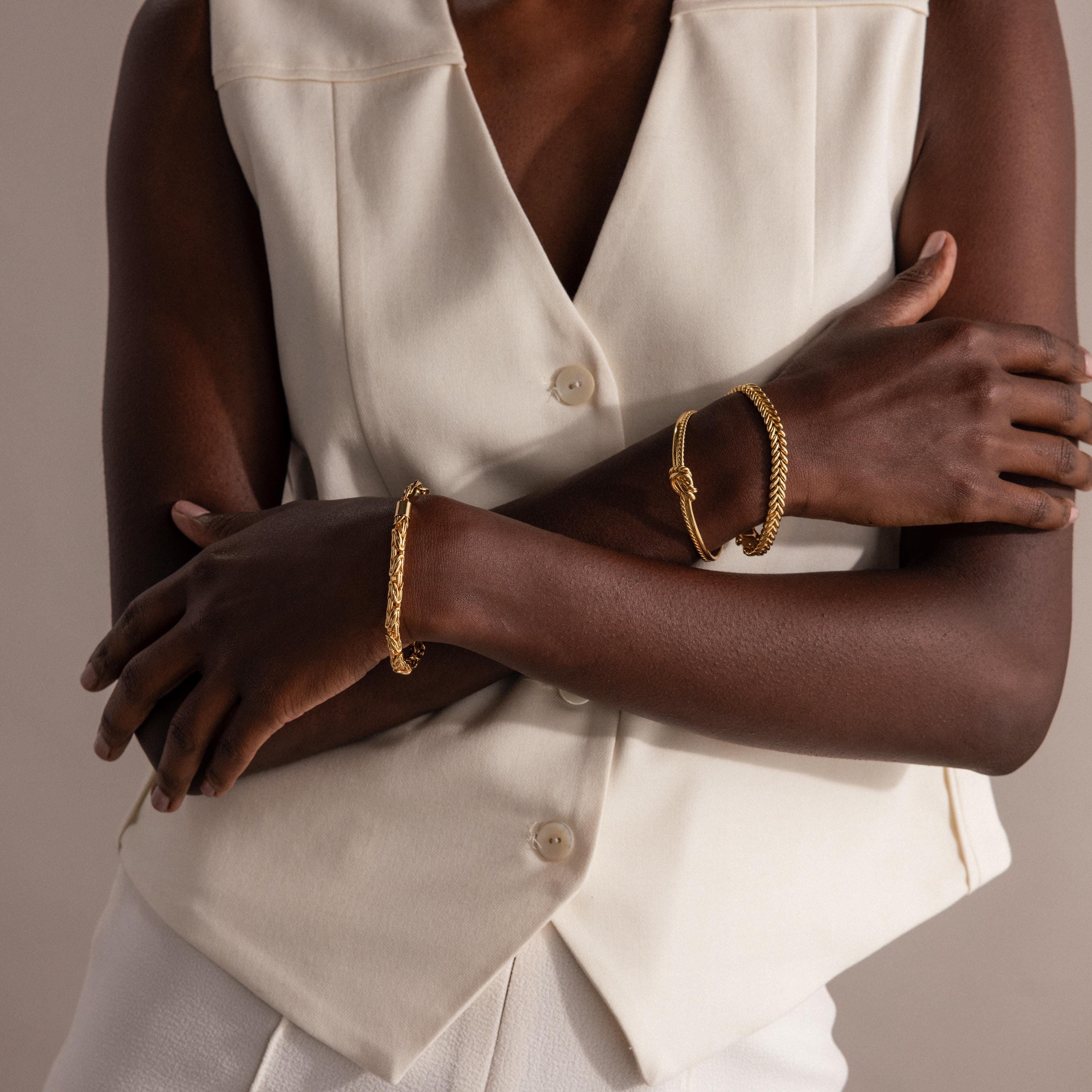 A woman in a cream vest and pants wears two gold Byzantine Link Bracelets, arms crossed, set against a neutral background.