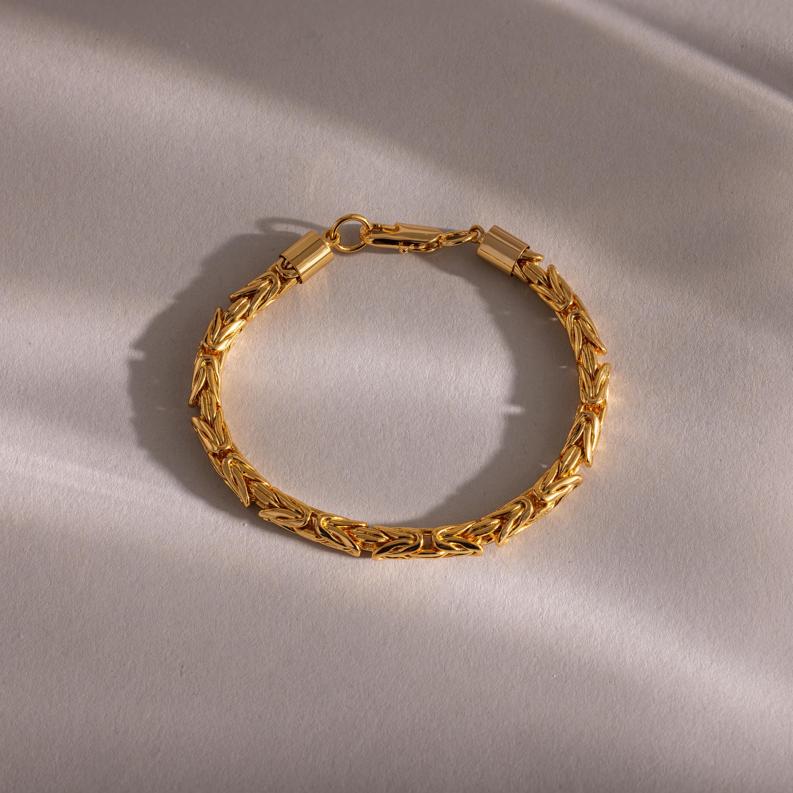 The Byzantine Link Bracelet features an intricate interwoven gold pattern, highlighted on a light textured surface with soft shadows, making it a striking statement piece.