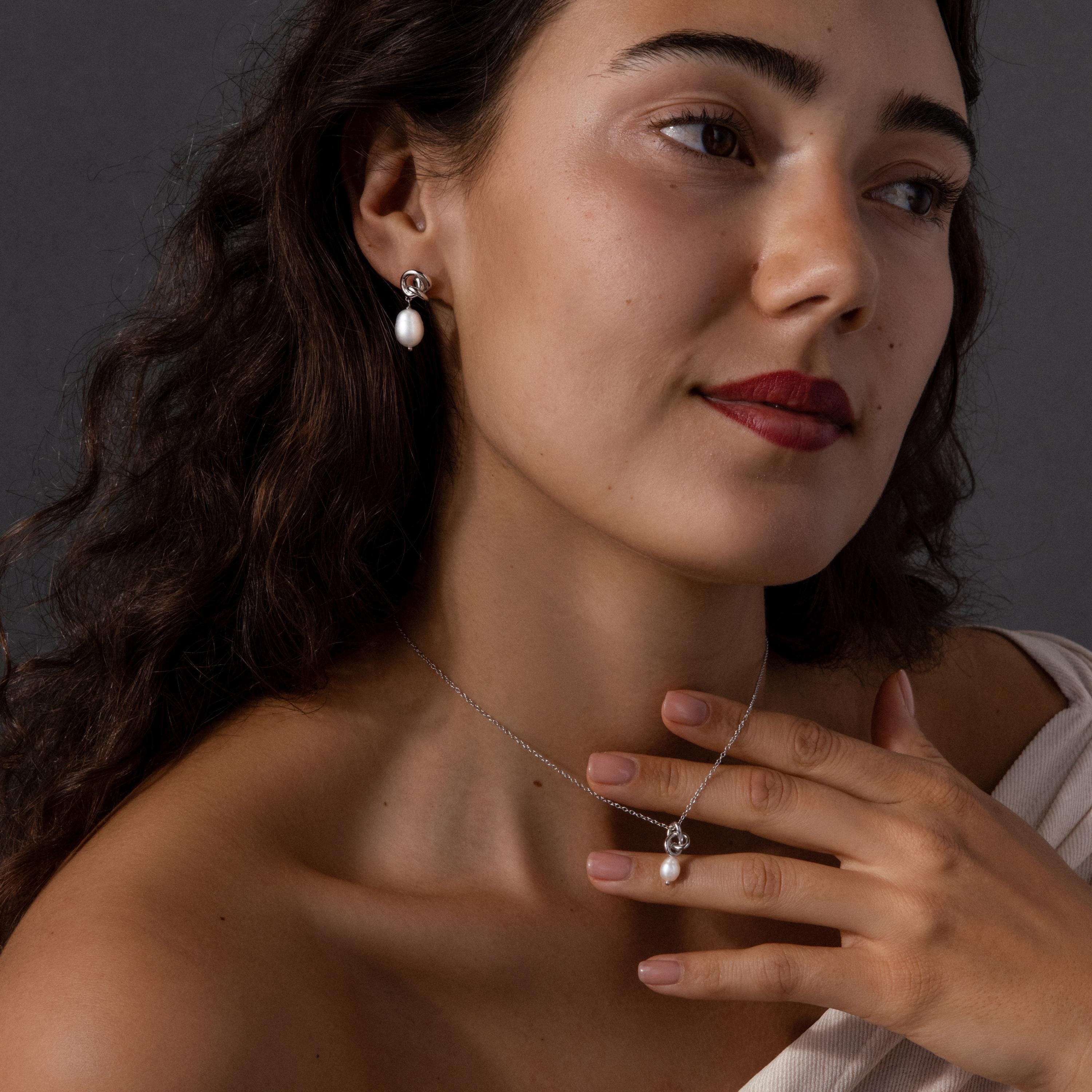 Woman with wavy hair wearing the Pearl Knot Necklace & Earrings Set in Sterling Silver, gently touching her neck on a dark background—an elegant choice for wedding jewelry.