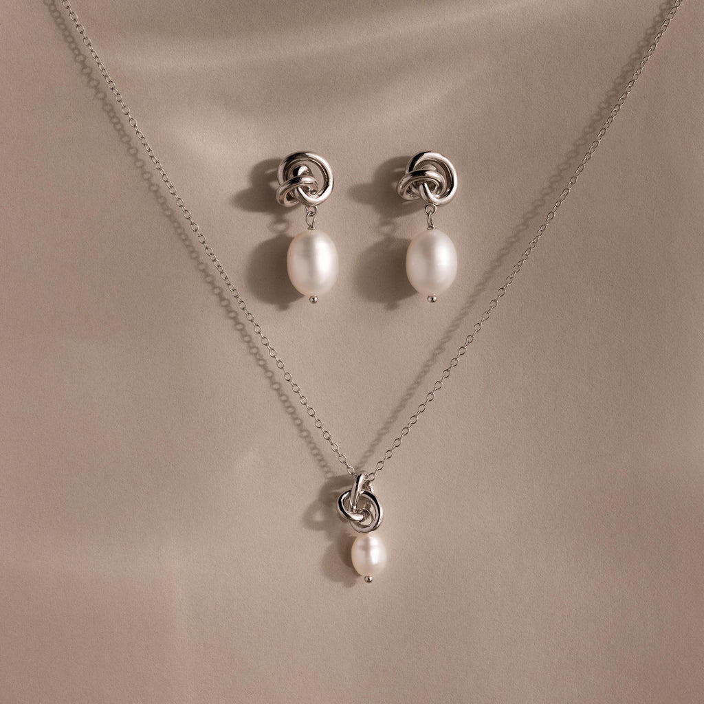 The Pearl Knot Necklace & Earrings Set in Sterling Silver features a delicate knot design with dangling white pearls, perfect for wedding jewelry and elegantly displayed on a beige surface.