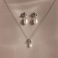 The Pearl Knot Necklace & Earrings Set in Sterling Silver features a delicate knot design with dangling white pearls, perfect for wedding jewelry and elegantly displayed on a beige surface.