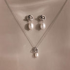 The Pearl Knot Necklace & Earrings Set in Sterling Silver features a delicate knot design with dangling white pearls, perfect for wedding jewelry and elegantly displayed on a beige surface.