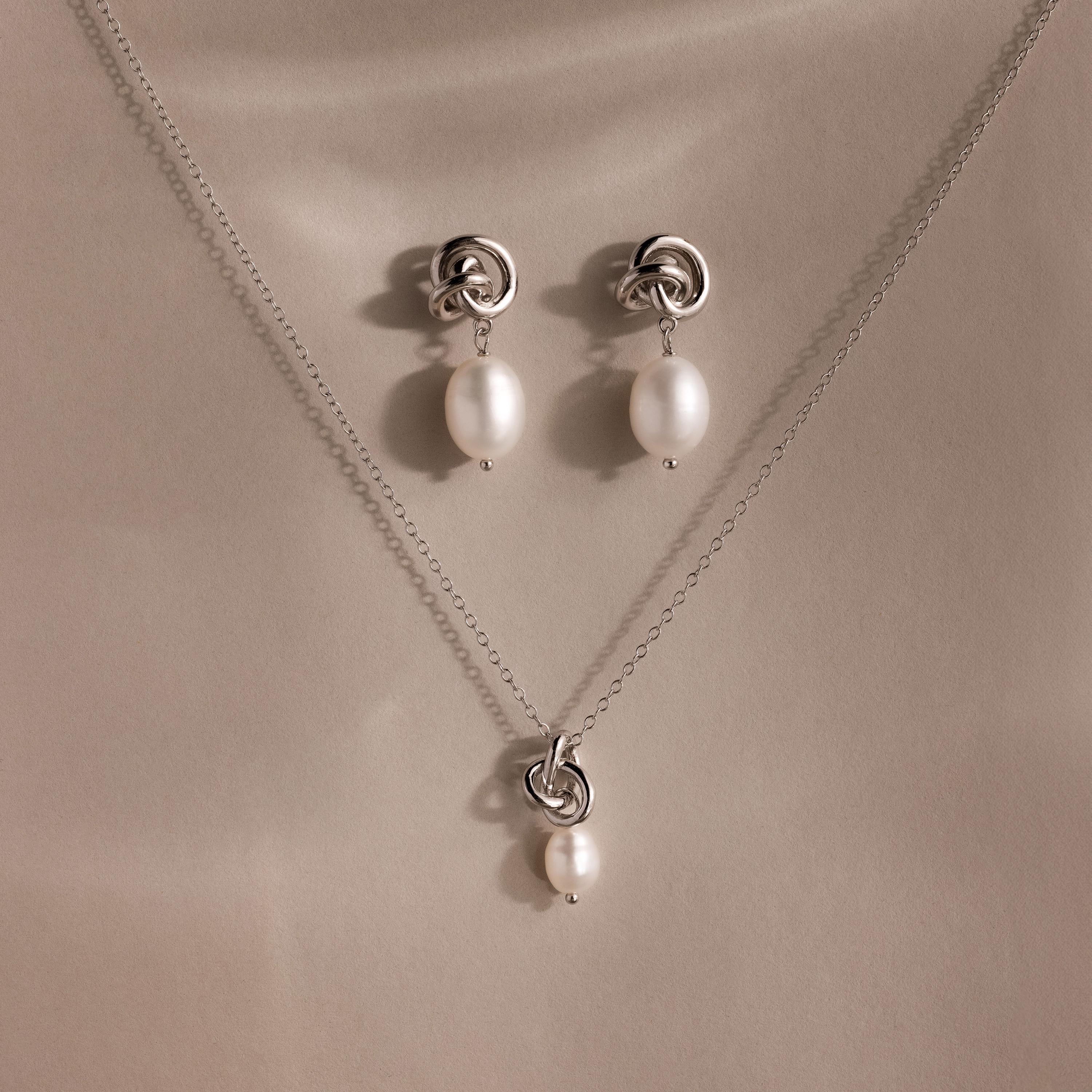 The Pearl Knot Necklace & Earrings Set in Sterling Silver features a delicate knot design with dangling white pearls, perfect for wedding jewelry and elegantly displayed on a beige surface.