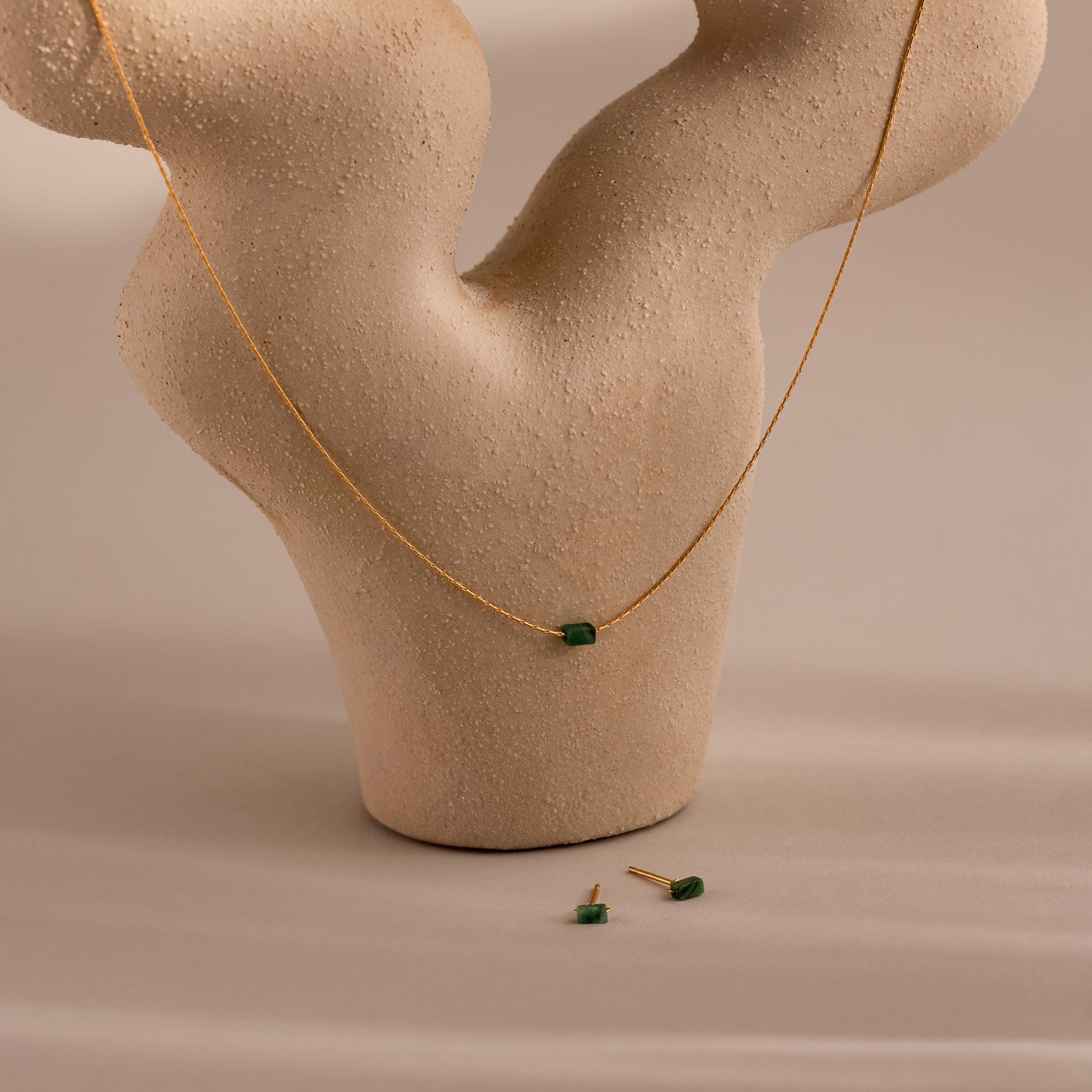 The Jade Bar Jewelry Set, featuring a gold necklace with a green jade stone and matching stud earrings, is displayed on a beige abstract textured stand.