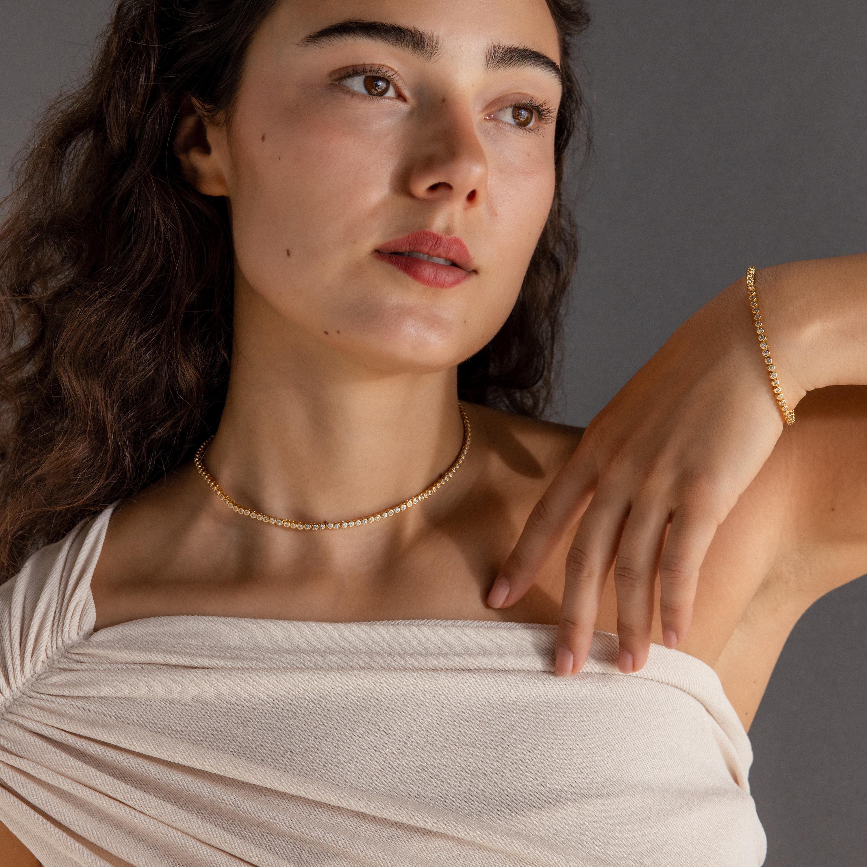 A woman in an off-shoulder cream top against a gray background wears a gold beaded necklace layered with the Diamond Bezel Tennis Necklace & Bracelet Set.