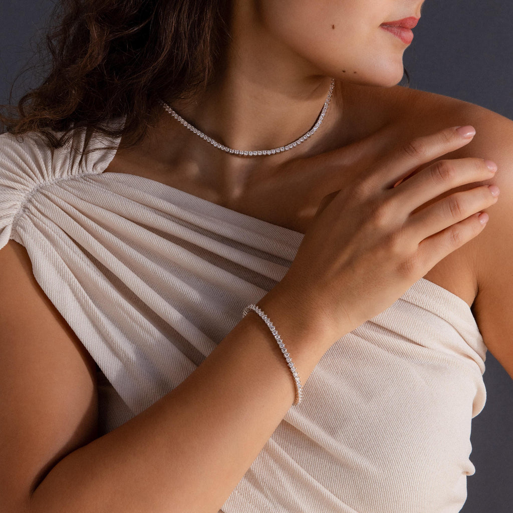 Woman in a cream, off-shoulder dress wears the 3-Prong Diamond Tennis Necklace & Bracelet Set in White Gold, gently touching her collarbone.