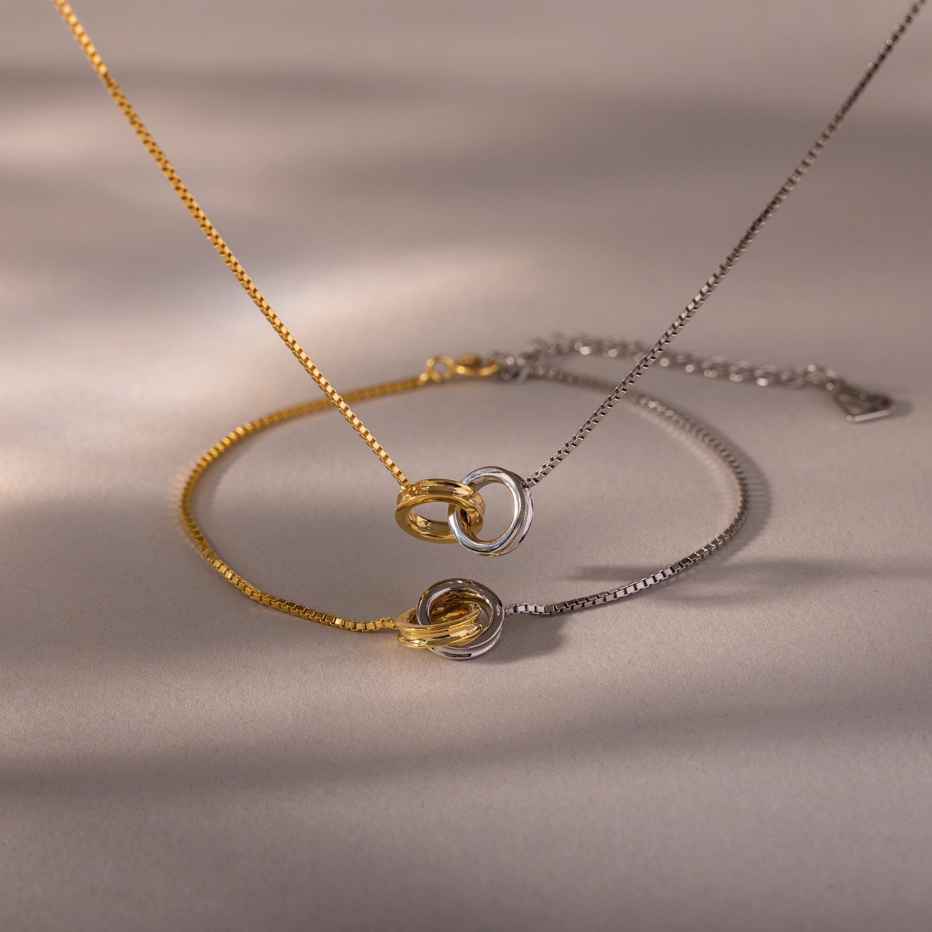 The Interlocking Circles Necklace and Bracelet Set in gold and silver, featuring minimalist design with interlocking rings, is displayed on a neutral surface with soft shadows.