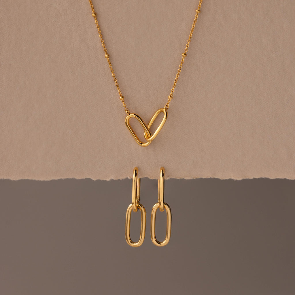 The Linked Charm Necklace and Earrings Set features two interlocking heart-shaped links and matching gold earrings, making an elegant anniversary gift displayed on a beige background.