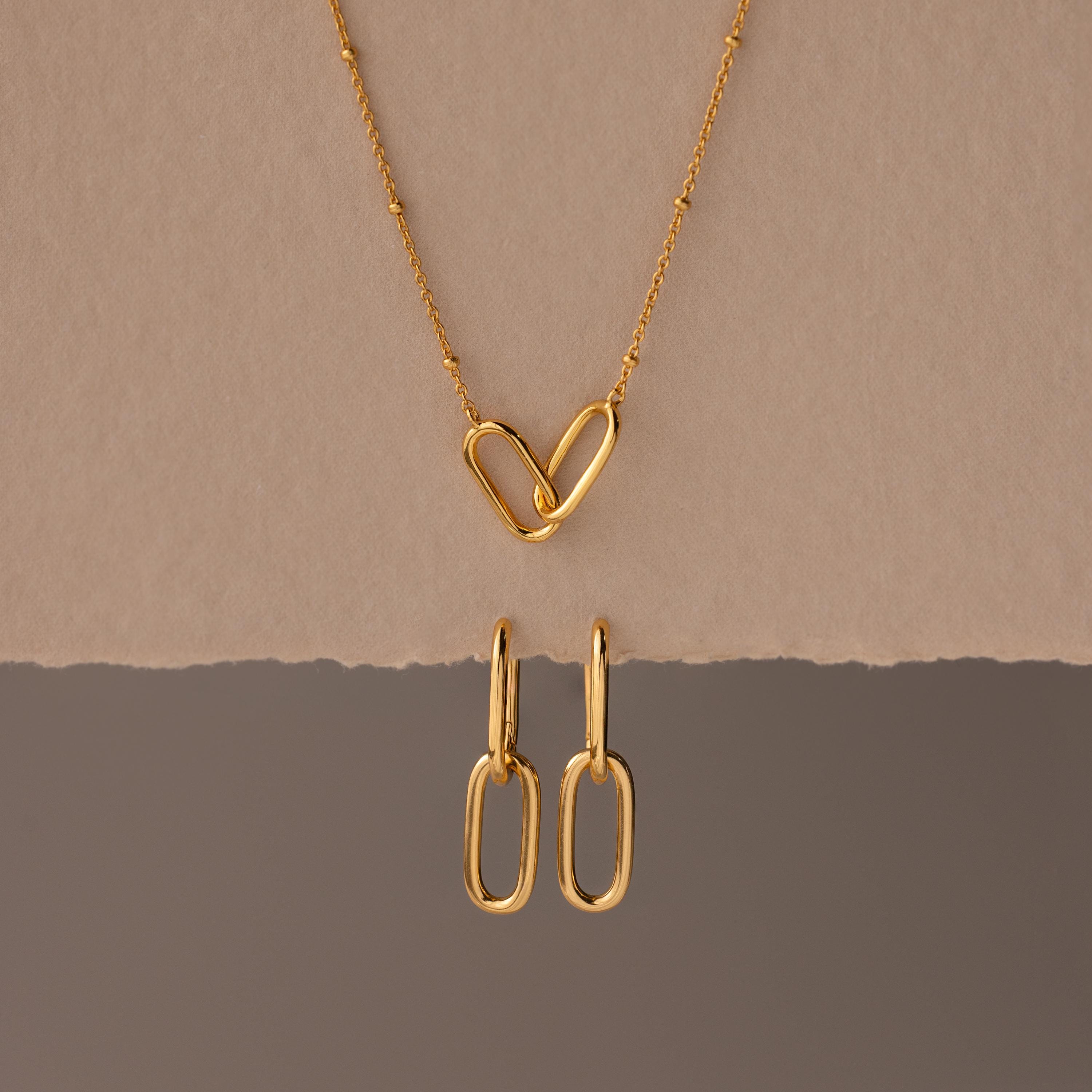 The Linked Charm Necklace and Earrings Set features two interlocking heart-shaped links and matching gold earrings, making an elegant anniversary gift displayed on a beige background.