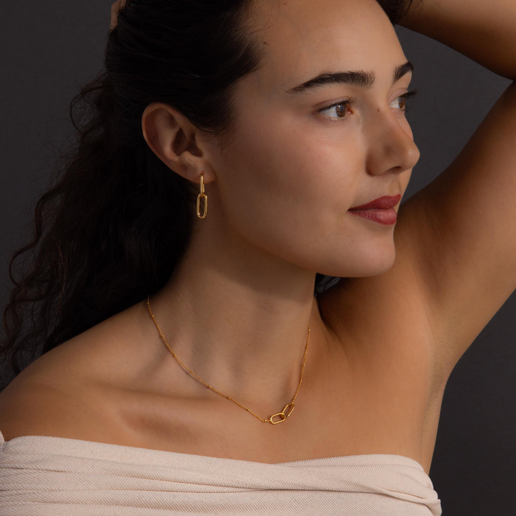 Woman with dark hair wears the Linked Charm Necklace and Earrings Set in 18K Gold, looking to the side against a dark background—an elegant choice for an anniversary gift.