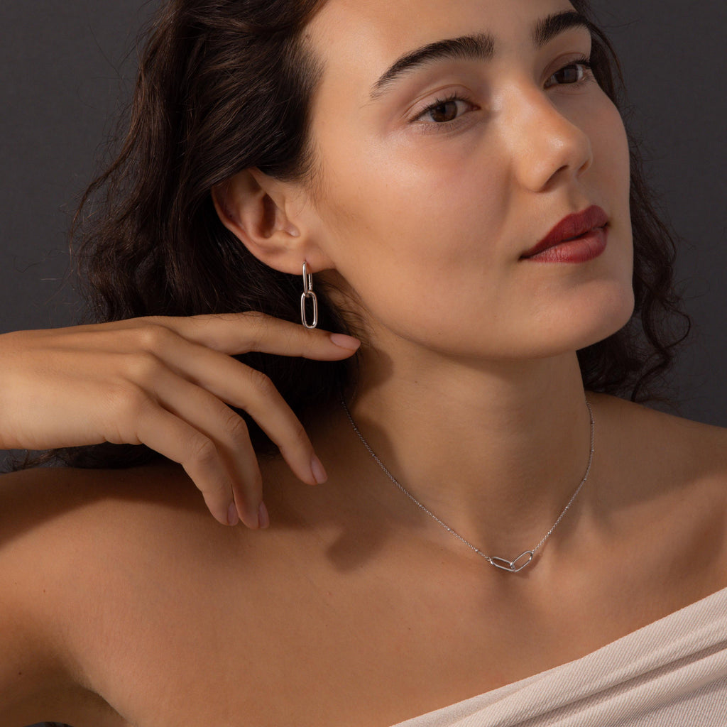 A woman poses against a dark background, gently touching her face while wearing the Linked Charm Necklace and Earrings Set in Sterling Silver—an elegant look ideal as an anniversary gift.