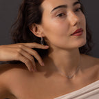 A woman poses against a dark background, gently touching her face while wearing the Linked Charm Necklace and Earrings Set in Sterling Silver—an elegant look ideal as an anniversary gift.