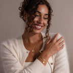 Smiling woman with curly hair and gold jewelry in a cream sweater touches her shoulder, showcasing the Fingerprint Birthstone Teardrop Necklace.