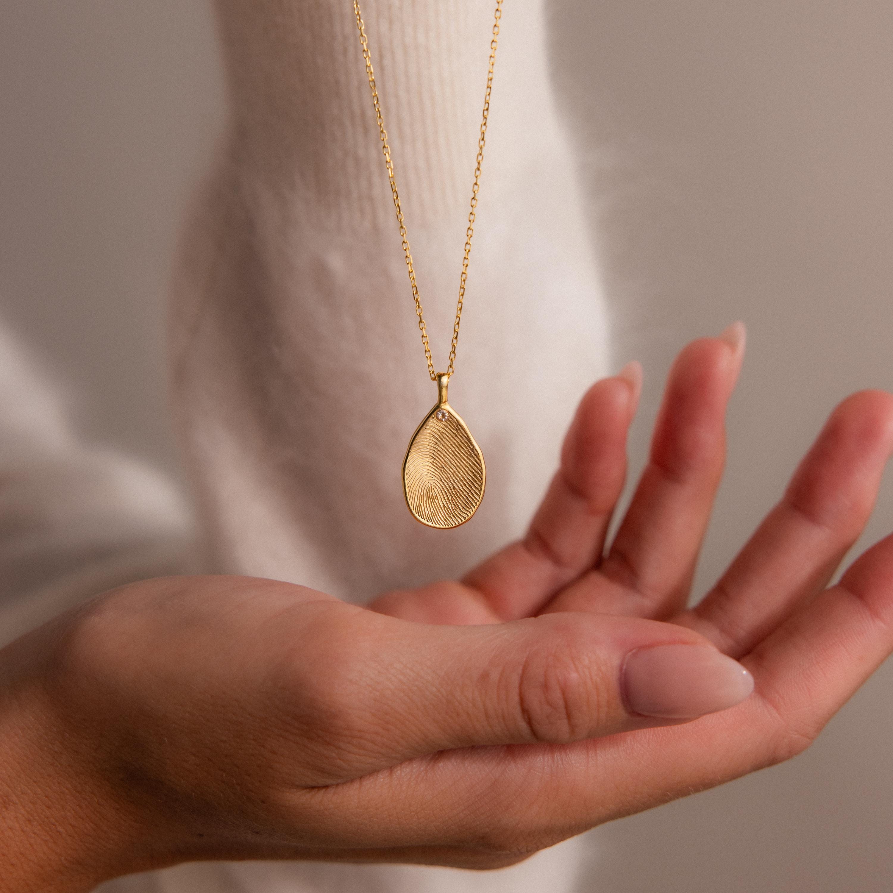 A hand holds the Fingerprint Birthstone Teardrop Necklace, a personalized gold pendant featuring a fingerprint and birthstone—perfect for capturing unique moments.