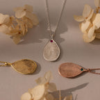 The Fingerprint Birthstone Teardrop Necklace in Sterling Silver rests on a beige surface, surrounded by dried flowers—a stunning personalized keepsake and unique jewelry piece.