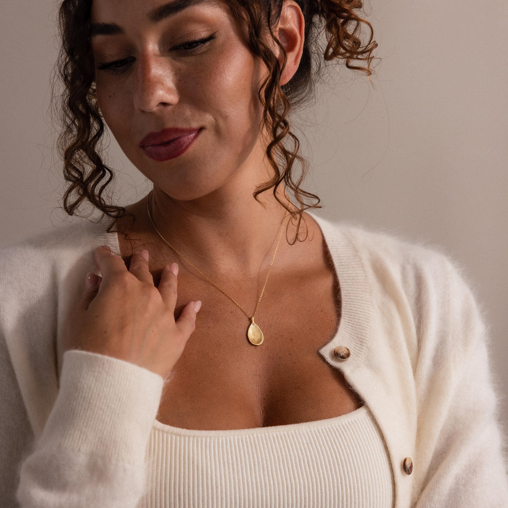 A woman with curly hair wears a white sweater and the Fingerprint Birthstone Teardrop Necklace in 18K Gold, looking down with a gentle smile, showcasing her unique custom keepsake.