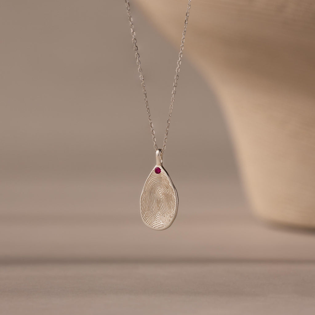 The Fingerprint Birthstone Teardrop Necklace features a teardrop silver pendant with your fingerprint and a red birthstone on a delicate chain—a personalized jewelry piece for meaningful style.