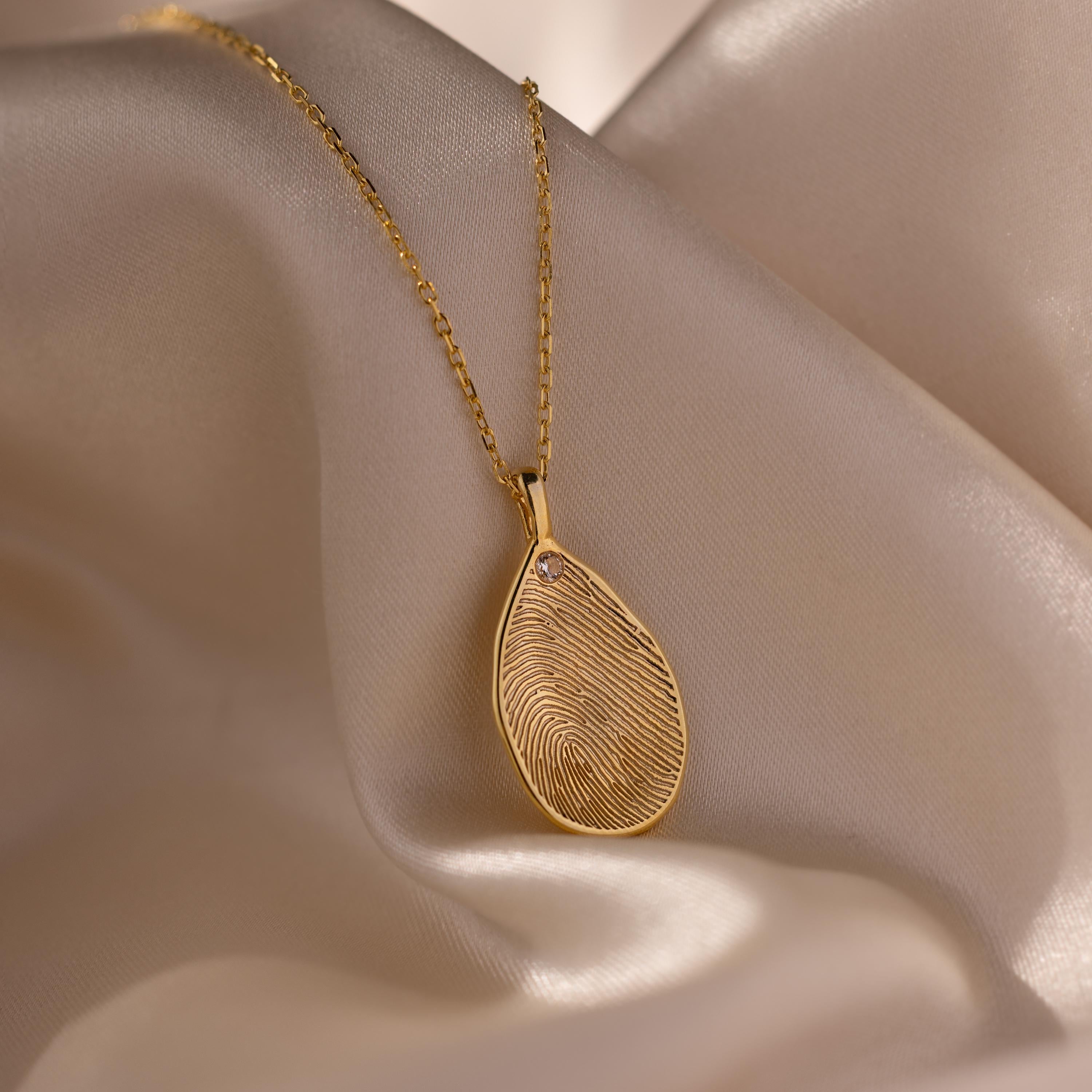 The Fingerprint Birthstone Teardrop Necklace in 18K Gold, custom engraved for a unique keepsake, is displayed on soft beige satin fabric.