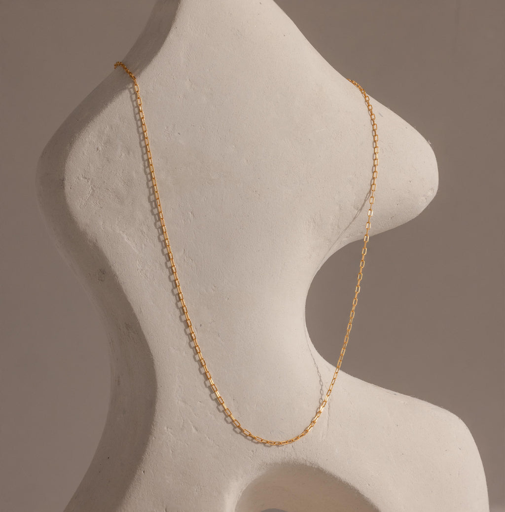 The Dainty Paperclip Necklace in 18K Gold drapes elegantly over a curvy white abstract sculpture, highlighting the modern sophistication of this delicate jewelry piece.