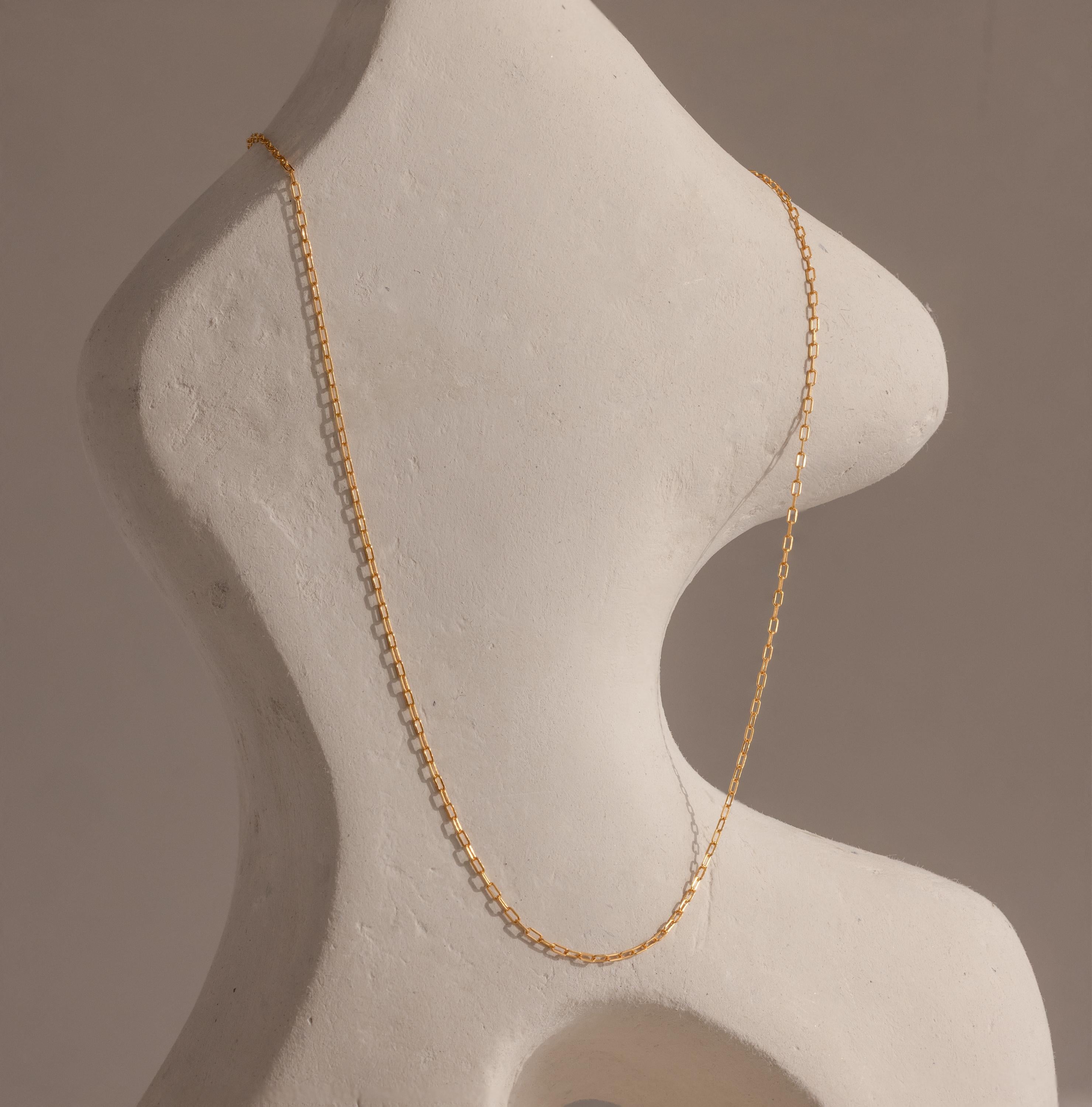 The Dainty Paperclip Necklace in 18K Gold drapes elegantly over a curvy white abstract sculpture, highlighting the modern sophistication of this delicate jewelry piece.