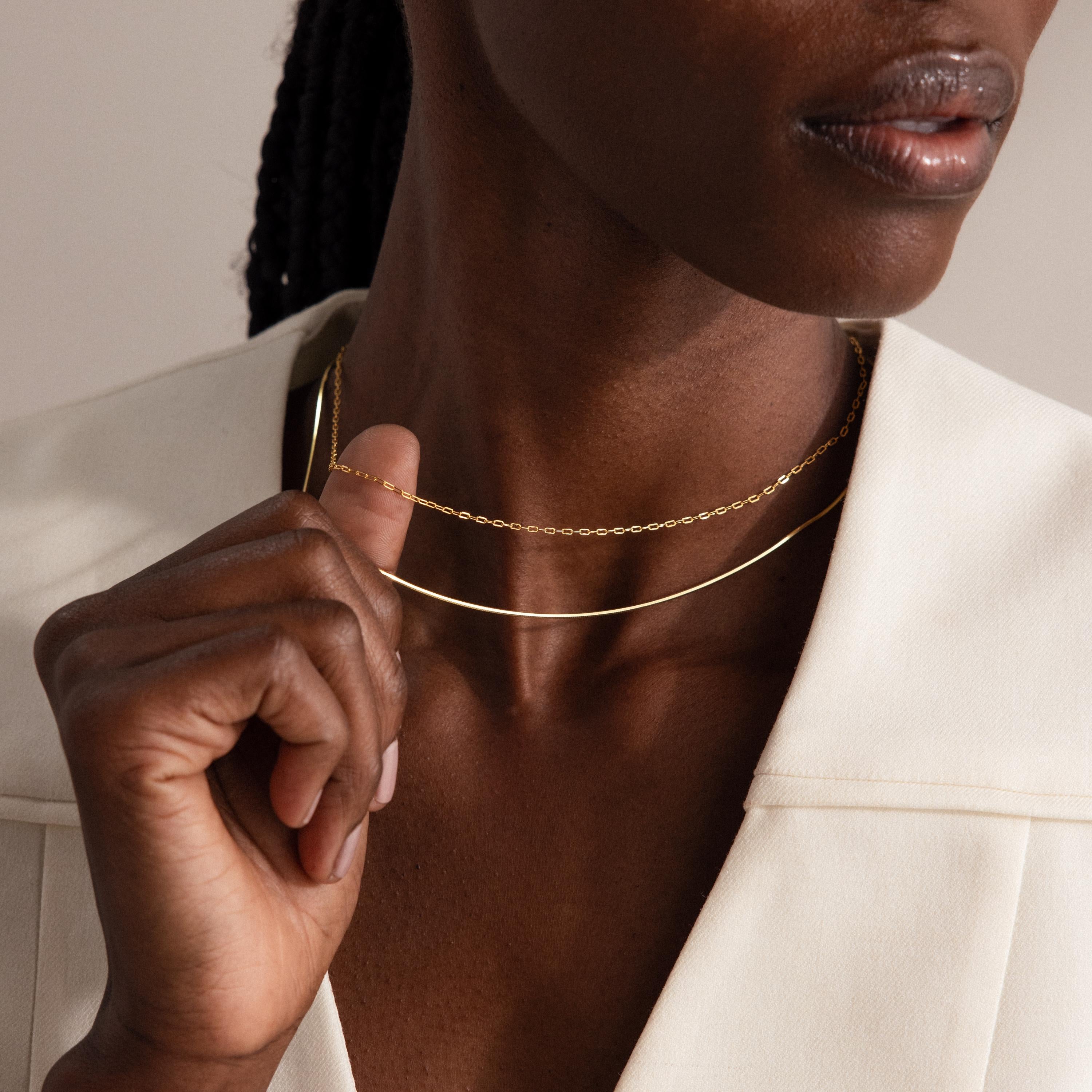 A woman in a cream top gently holds the Dainty Paperclip Necklace in 18K Gold, styled with another delicate gold necklace.