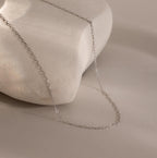 The Dainty Paperclip Necklace features a delicate silver chain, elegantly draped over a smooth white stone—perfect for layering and adding subtle shine against any neutral background.