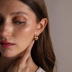 A woman with long brown hair gently touches her Bold Teardrop Earrings in 18K Gold, which feature a high polish finish.