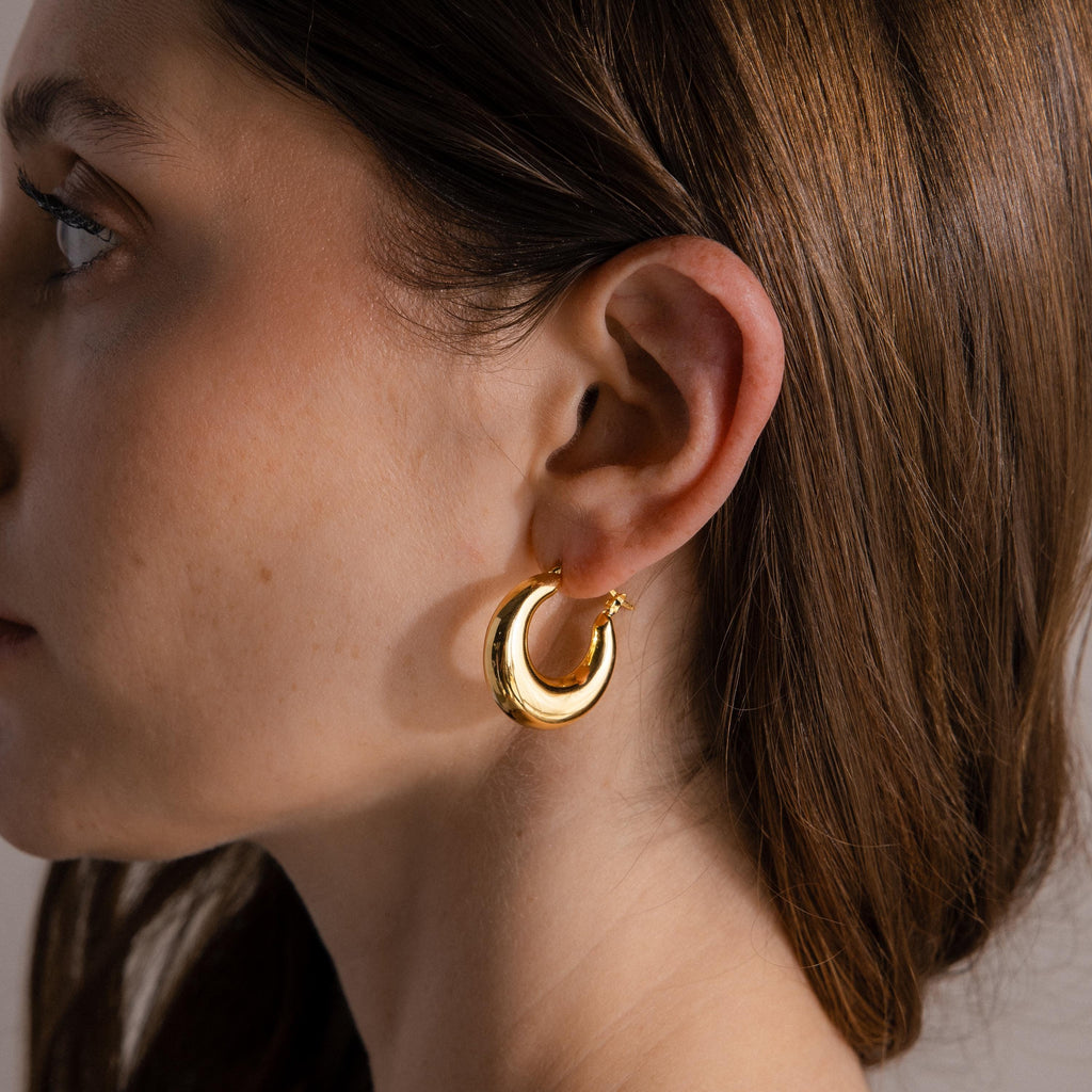 Close-up of a woman’s ear wearing Sculpted Hoops in 18K Gold, shiny gold hoop earrings radiating timeless elegance, with brown hair tucked behind her ear.