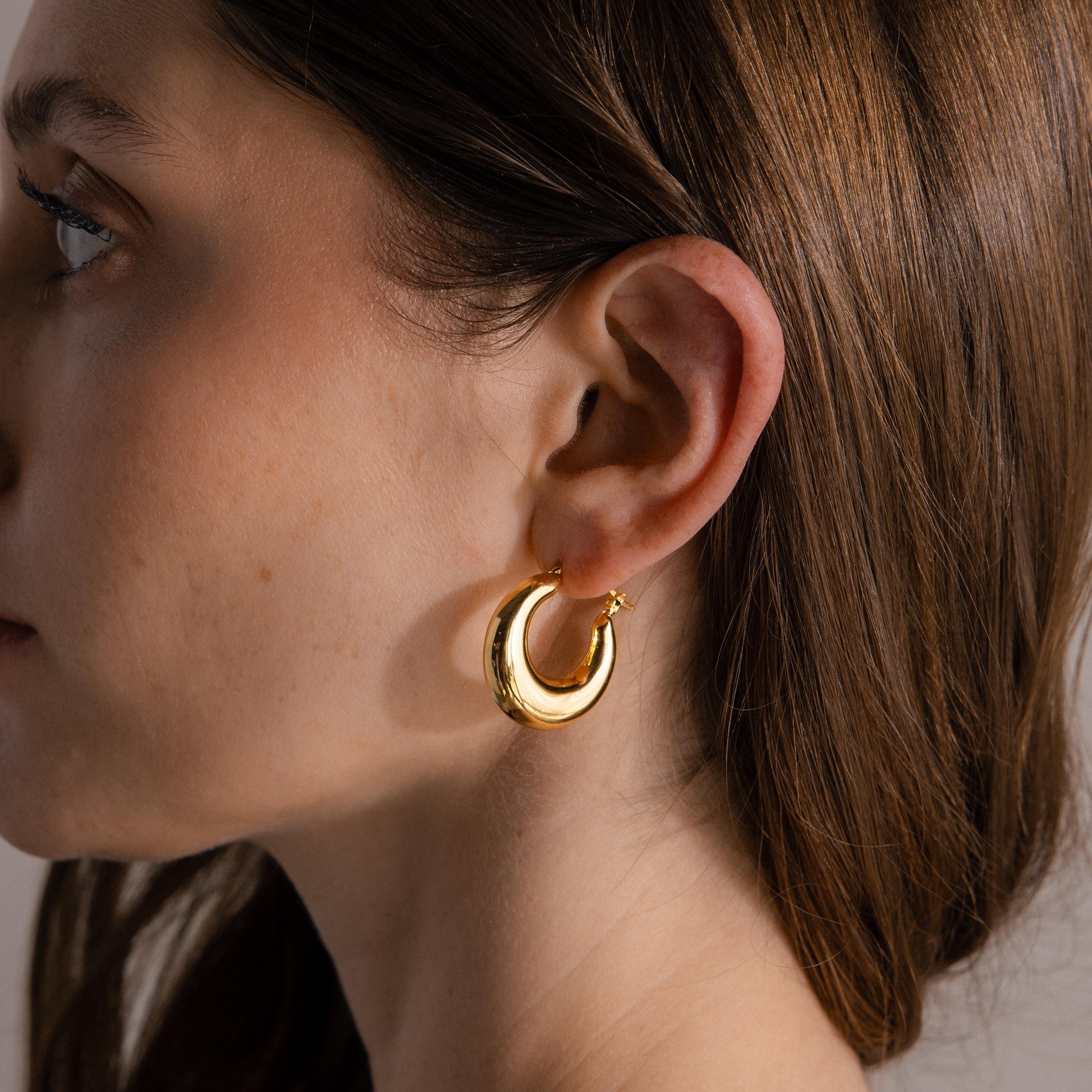 Close-up of a woman’s ear wearing Sculpted Hoops in 18K Gold, shiny gold hoop earrings radiating timeless elegance, with brown hair tucked behind her ear.