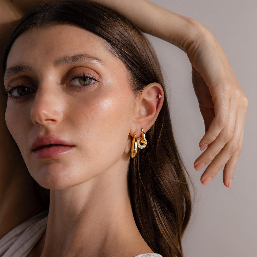 A woman with long brown hair wears Sculpted Hoops earrings, looking at the camera with her hand raised near her head.