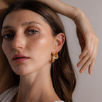 A woman with long brown hair wears Sculpted Hoops earrings, looking at the camera with her hand raised near her head.