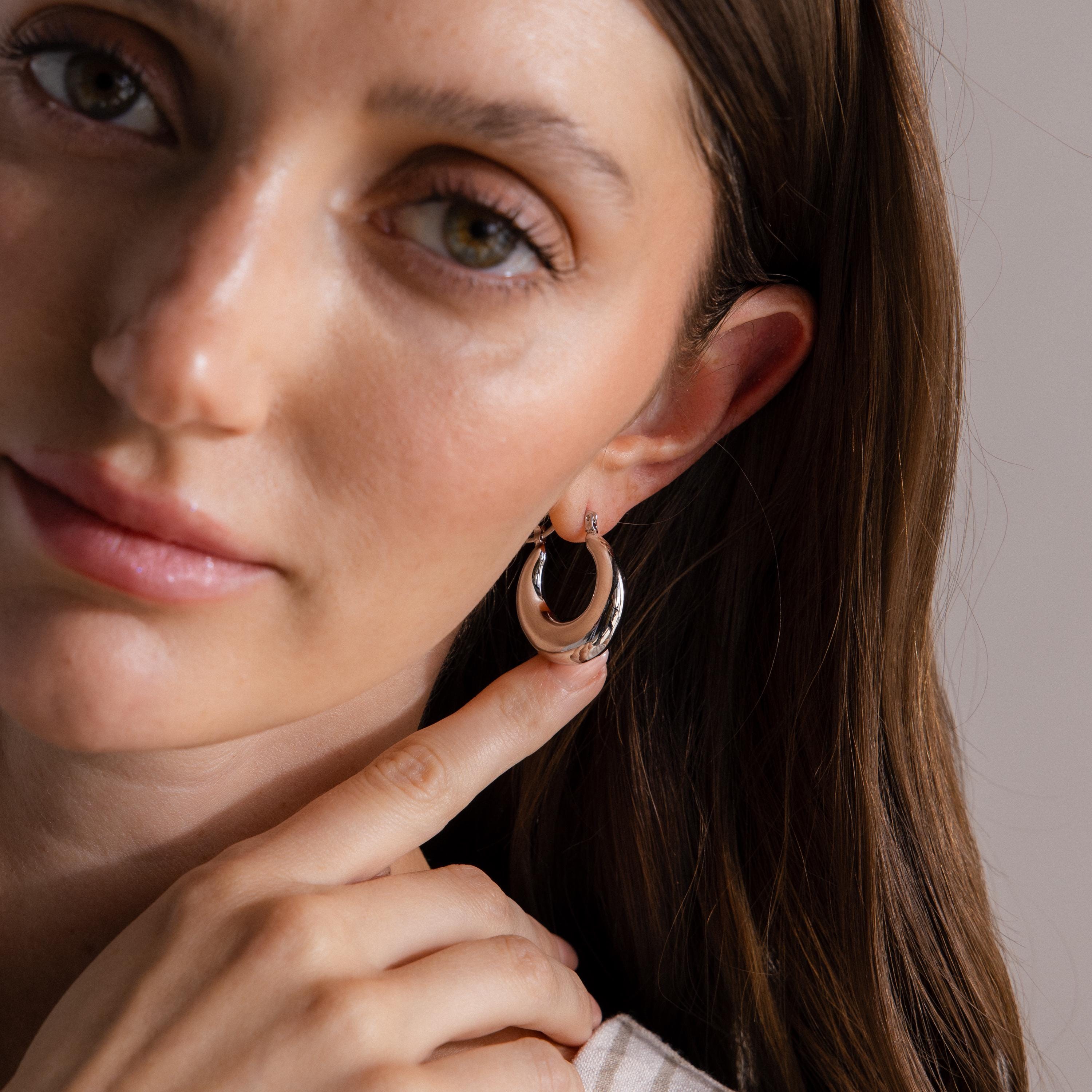 A woman with long brown hair points to her ear, showcasing the high shine finish of Sculpted Hoops, large silver earrings.