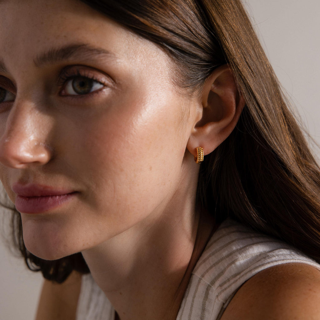 Woman with long brown hair gazes to the side in soft lighting, wearing Beaded Chunky Hoops in 18K Gold.