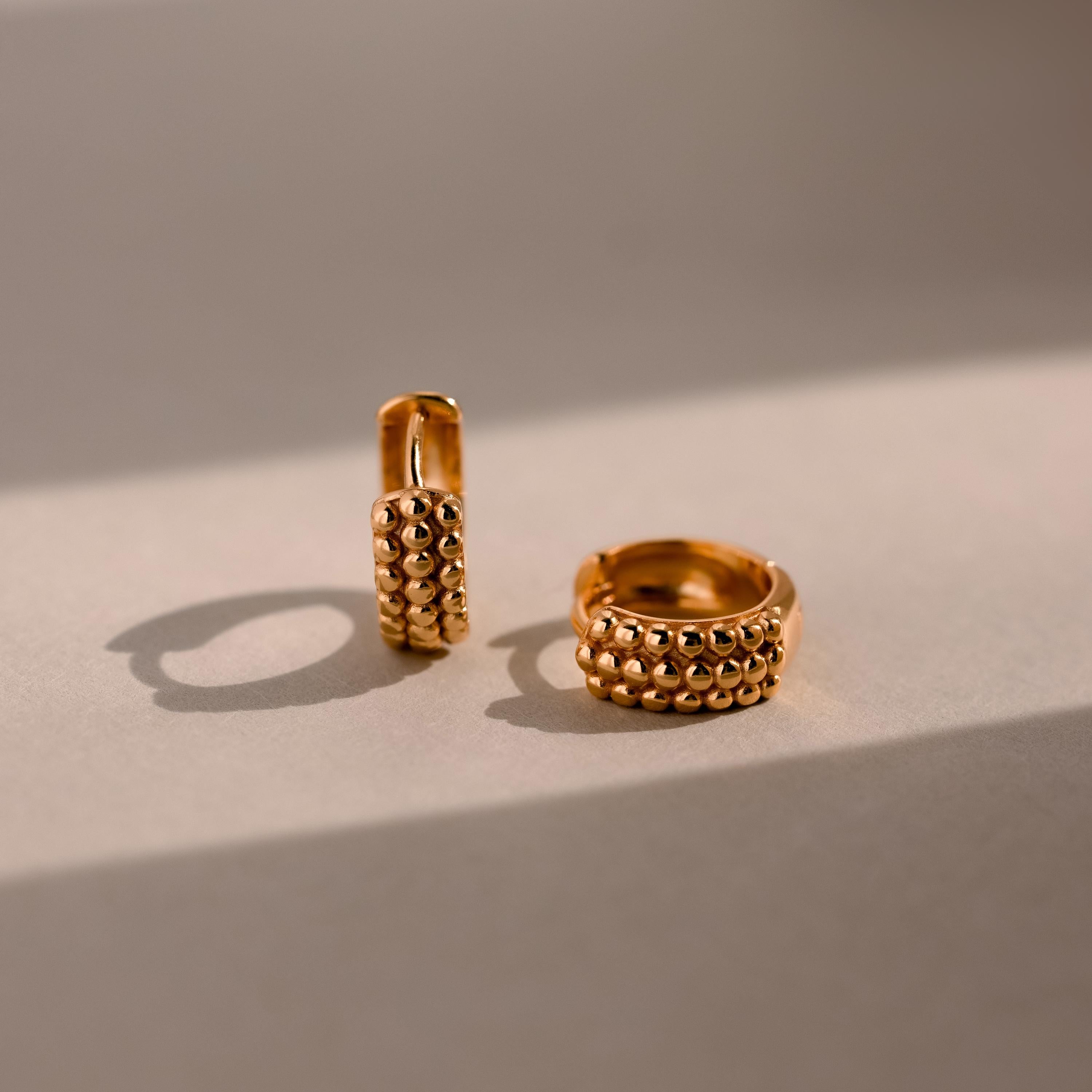 Two Beaded Chunky Hoops in 18K Gold rest on a sunlit neutral surface, their textured design casting soft shadows and highlighting the earrings’ modern, elegant shimmer.