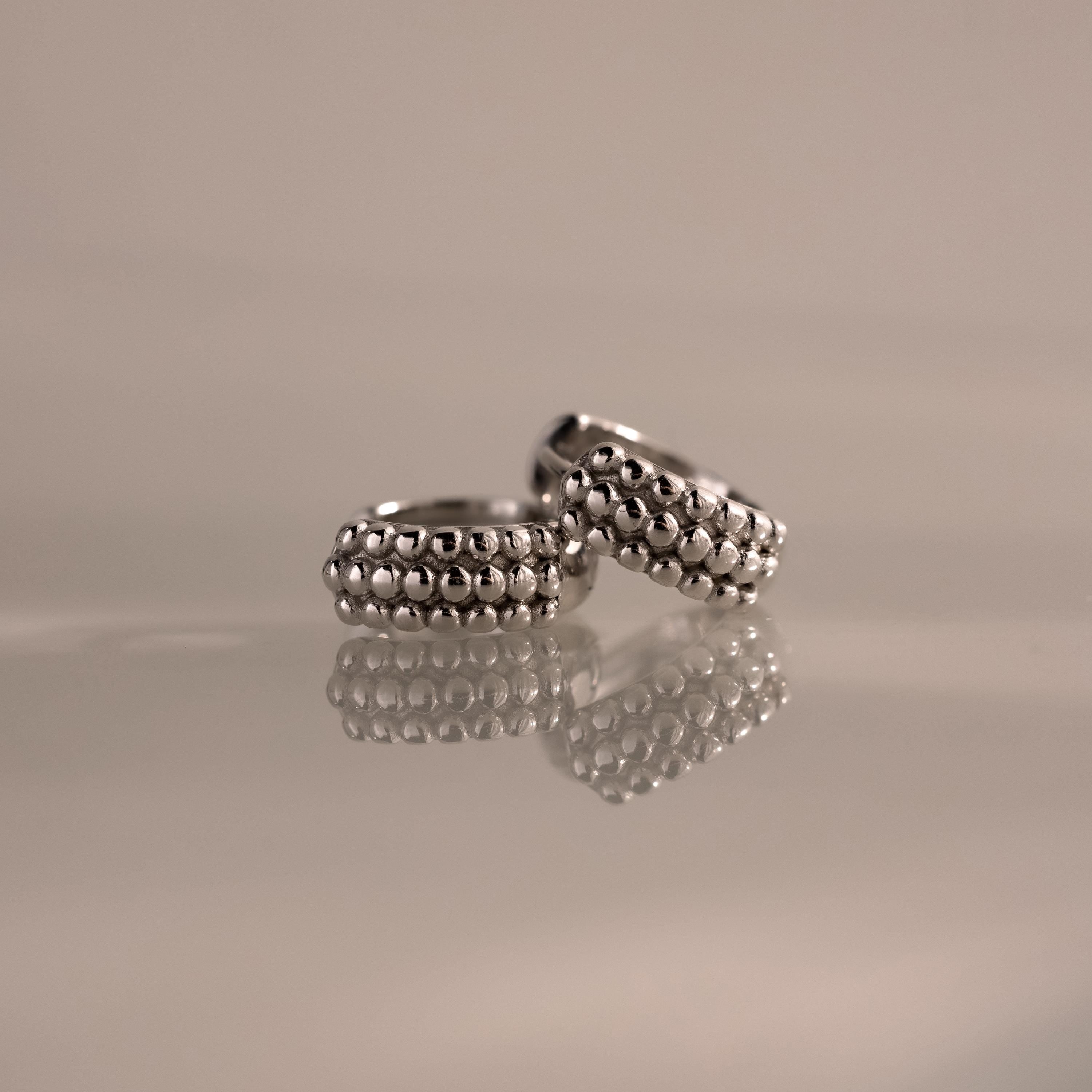 Beaded Chunky Hoops, two silver rings with a beaded texture inspired by classic styles, are reflected on a glossy surface against a neutral background.