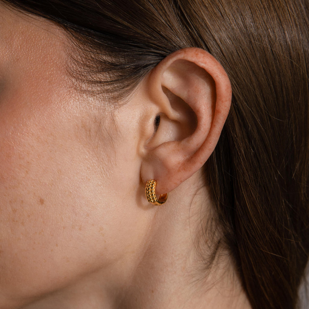Close-up of a woman's ear with straight brown hair, wearing Beaded Chunky Hoops—a small gold earring with a textured, beaded design for a modern edge.