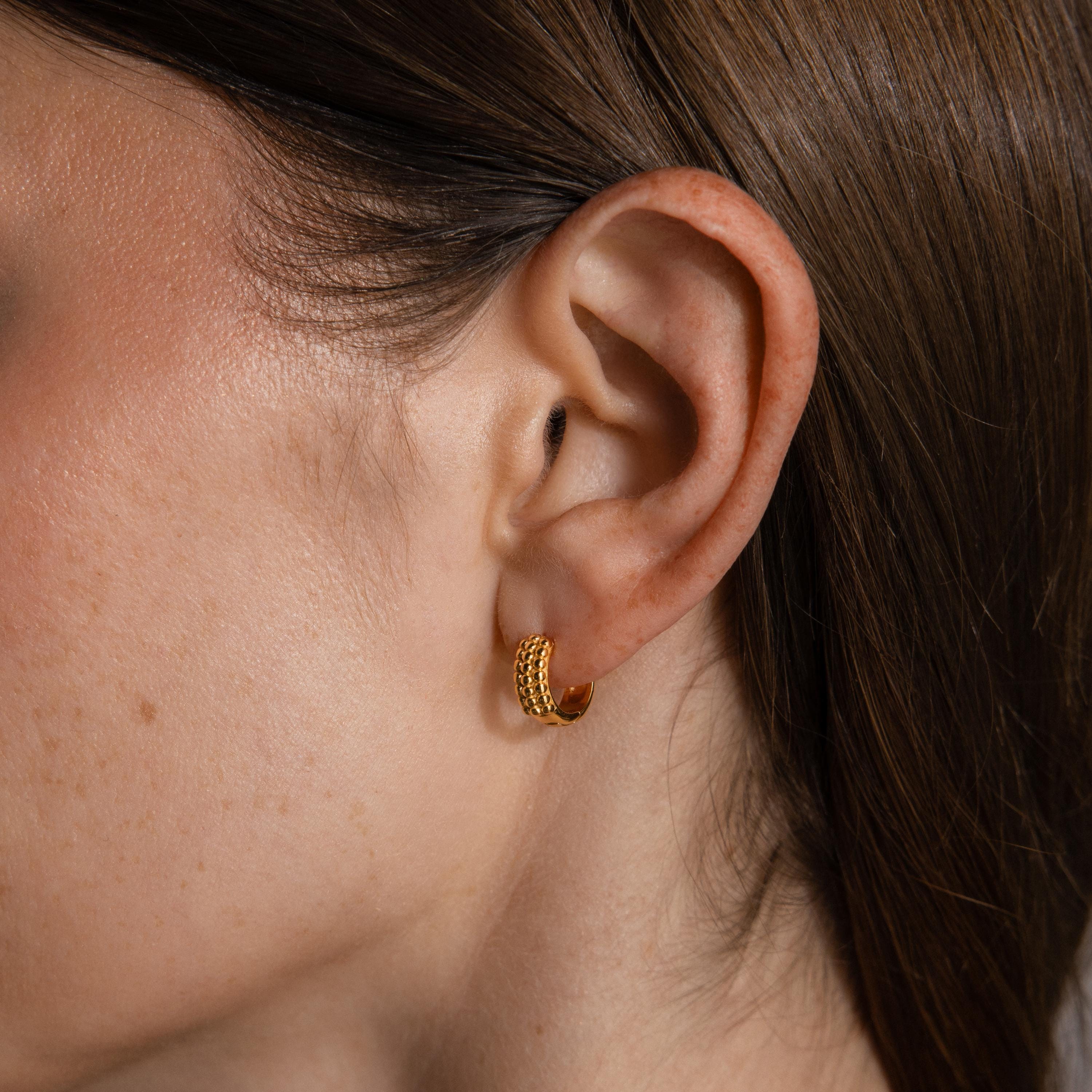 Close-up of a woman's ear with straight brown hair, wearing Beaded Chunky Hoops—a small gold earring with a textured, beaded design for a modern edge.