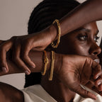 A woman with braided hair elegantly crosses her arms, showcasing three gold bracelets, including the eye-catching Byzantine Link Bracelet.