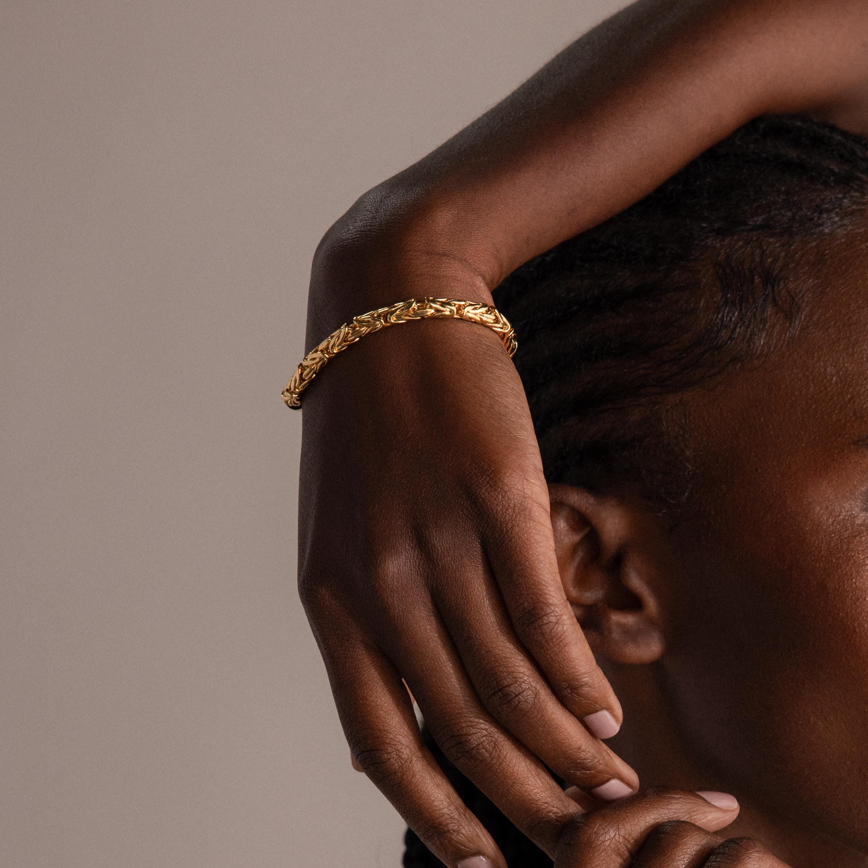 The Byzantine Link Bracelet in 18K Gold, with its elegant interwoven design, adorns a wrist resting near a forehead against a neutral background, making it a refined and subtle statement piece.