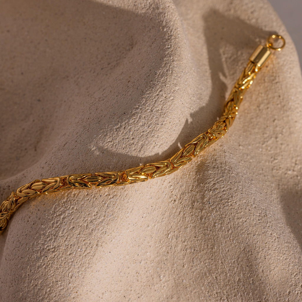 The Byzantine Link Bracelet in 18K Gold, featuring an intricate interwoven design, rests on a textured beige stone surface—a perfect statement jewelry piece.