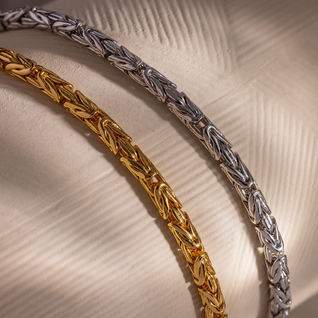 The Byzantine Link Bracelet features an intricate interwoven design, creating a stunning statement piece showcased on a textured beige surface.