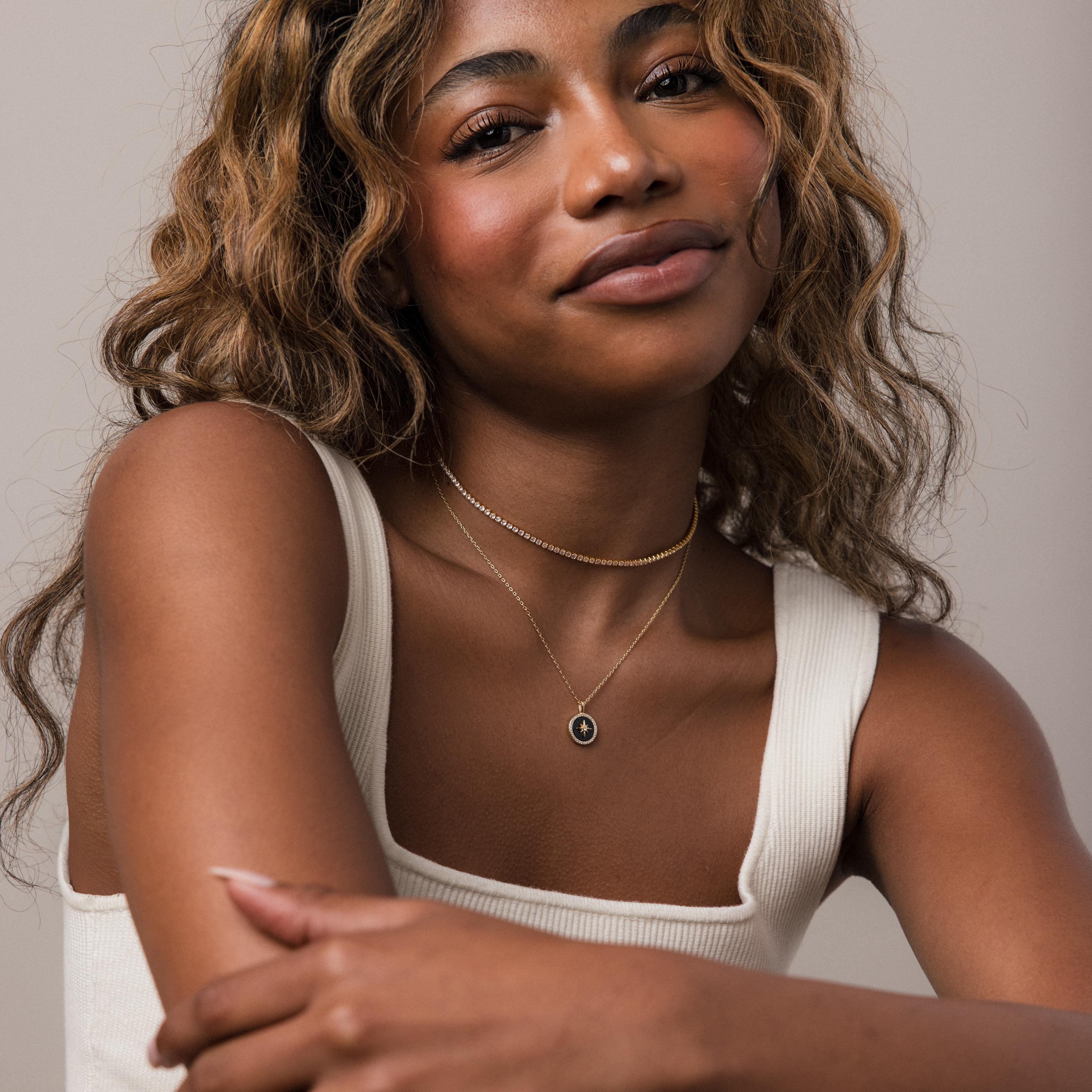 A woman with wavy hair in a white tank top smiles softly at the camera, wearing layered gold necklaces featuring the Onyx Star Pendant Necklace in 18K Gold.