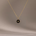 The Onyx Star Pendant Necklace in 18K Gold features a circular black onyx pendant with a star design, creating a striking celestial look against a neutral background.