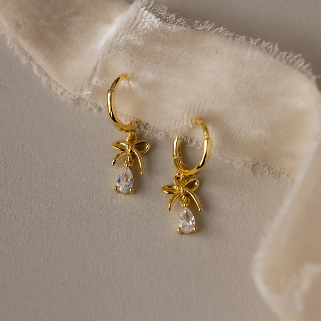 Flatlay of our Teardrop Ribbon Charm Hoops in Gold hoop earrings on a soft fabric background