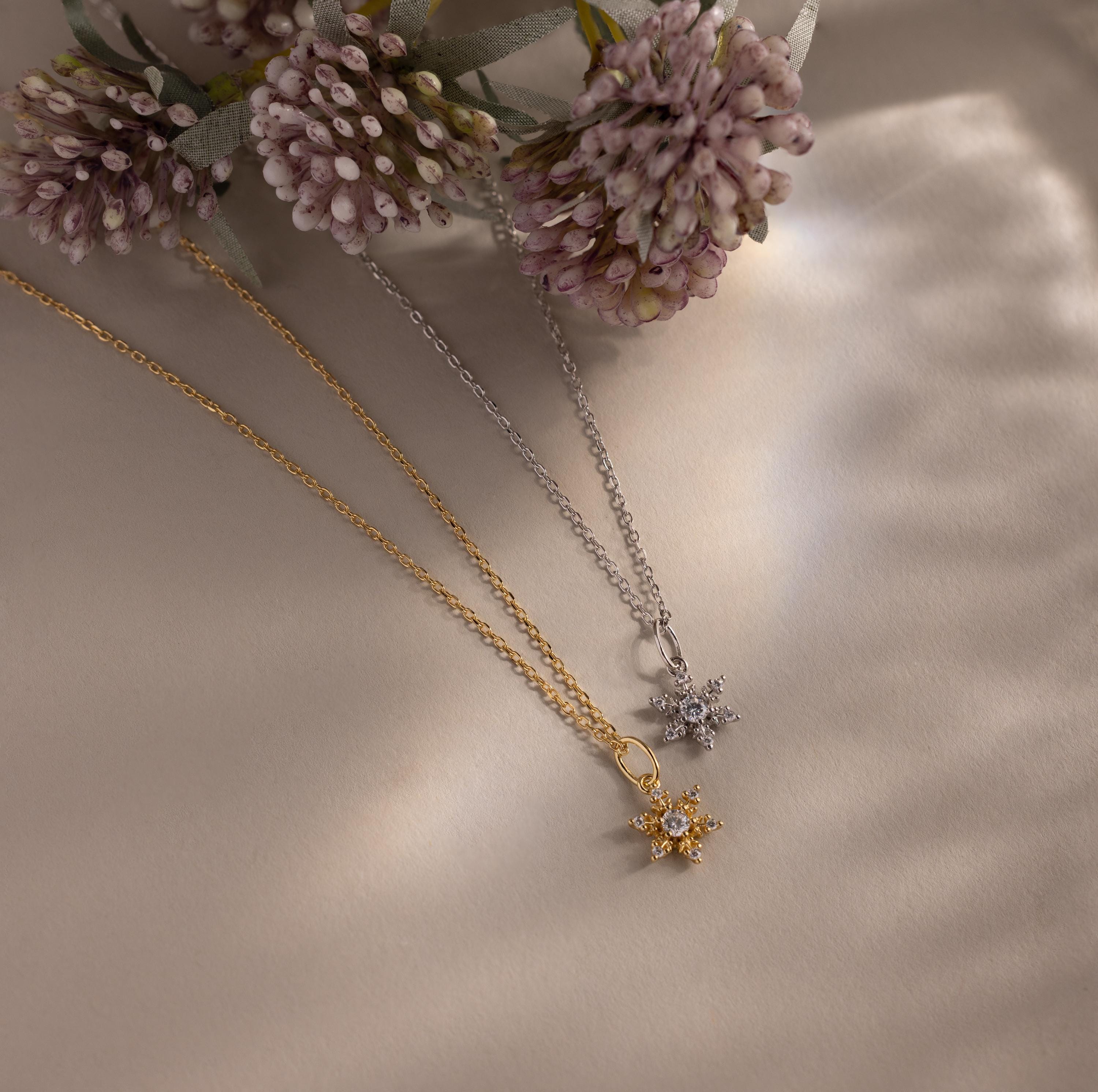 Flatlay of our Diamond Snowflake Necklace in Gold and Silver with light purple flowers in the background