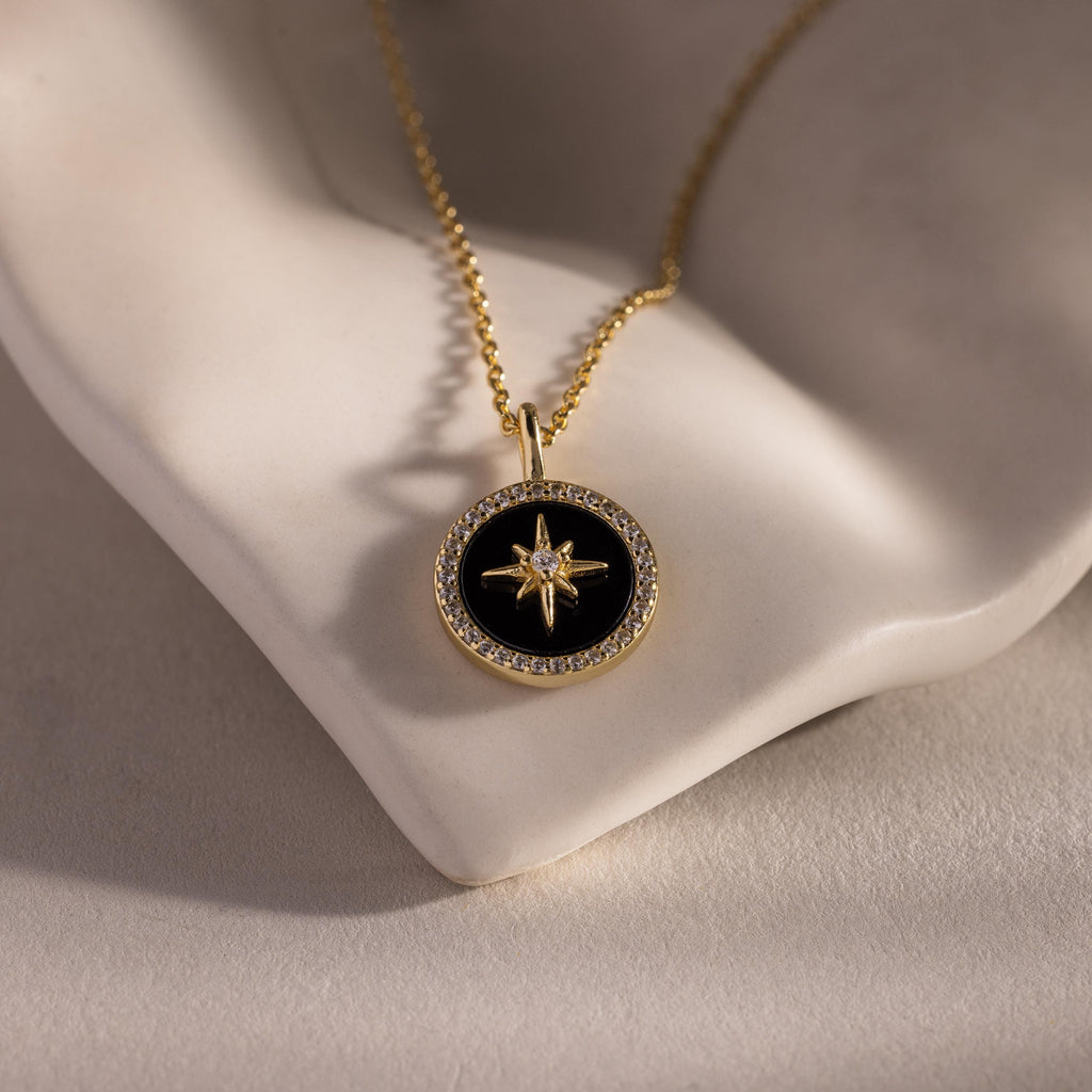 The Onyx Star Pendant Necklace in 18K Gold features a black onyx star and celestial design, displayed on a white sculpted surface. Ideal for anyone who loves elegant cosmic-inspired jewelry.