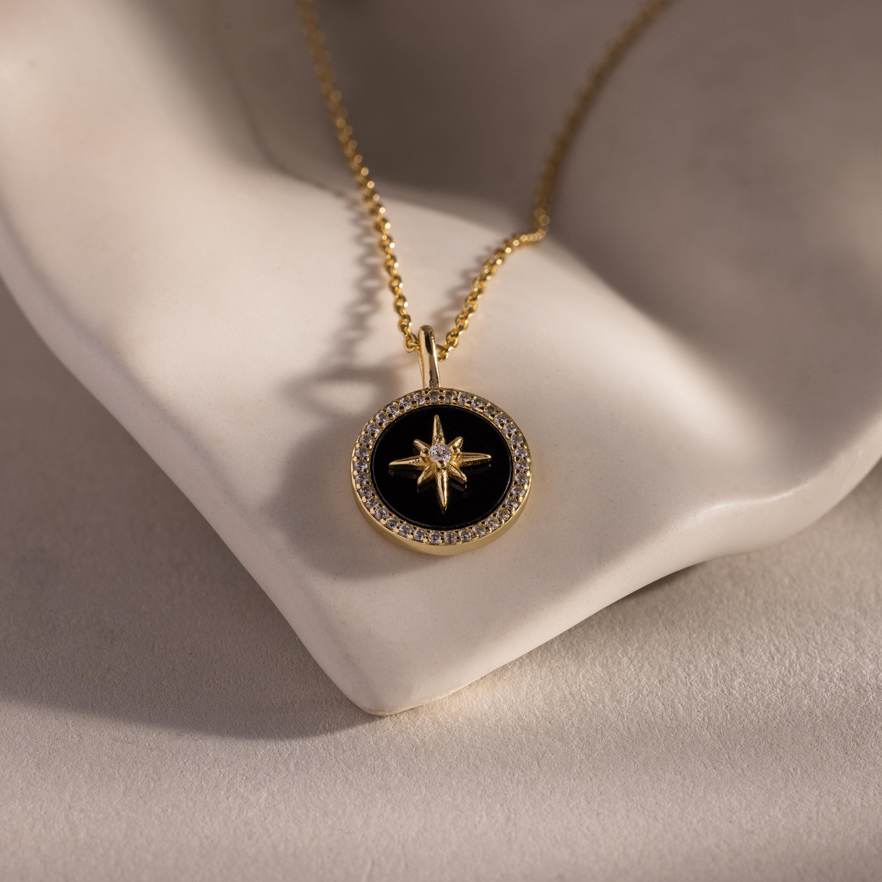 The Onyx Star Pendant Necklace in 18K Gold features a black onyx star and celestial design, displayed on a white sculpted surface. Ideal for anyone who loves elegant cosmic-inspired jewelry.